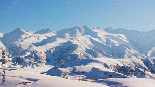Aerial view captures majestic snow-covered mountains in Kyrgyzstan under bright sunlight. Serene winter landscape showcases tranquility and natural beauty.