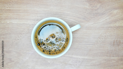Top-down view white ceramic cup filled with freshly brewed black coffee sitting on light wooden table.