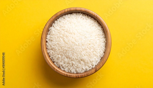 “Uncooked white rice in a wooden bowl on bright yellow background with scattered grains.”