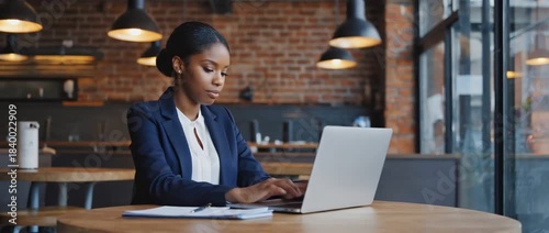 Focused Professional: A poised woman, deeply immersed in her work, sits at a table with her laptop, illuminated by the warm glow of the indoor setting, embodying diligence and dedication.