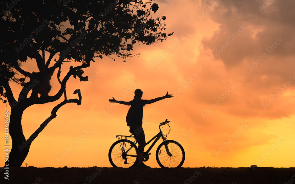 Obraz premium Silhouette of a person cycling at sunset with open arms under tree, freedom and outdoor lifestyle