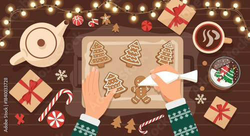 Hands decorating festive christmas gingerbread cookies with icing on a tray, surrounded by presents, candy canes, and a warm drink