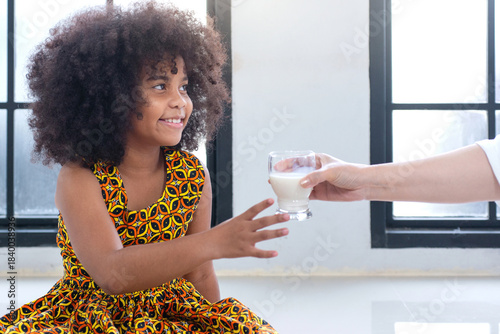 African child girl in traditional dress smiles and reaches out to receive a glass of milk from someone.