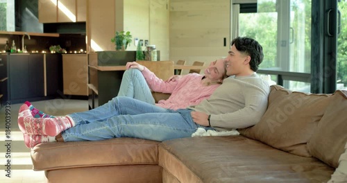 Diverse couple entering then settling onto sectional sofa in room, pointing at off-screen view