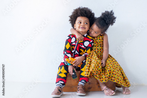 Boy and a girl, dressed in traditional African clothing, sit and playfully interact on a wooden box against a white background.