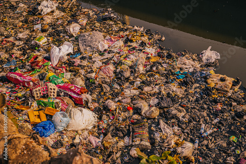 A polluted river flowing through the slums of Mumbai, filled with plastic waste, debris and untreated sewage. The scene shows densely built informal housing, overcrowded living conditions and the envi