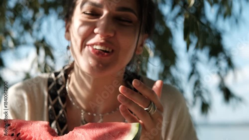 A beautiful brunette in a bohemian style relaxes outdoors in the summer, holding a large slice of watermelon against a green forest backdrop.