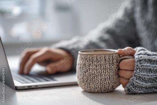 Woman in warm sweater holding coffee mug while working on laptop. Cozy office space for comfortable remote work or studying.