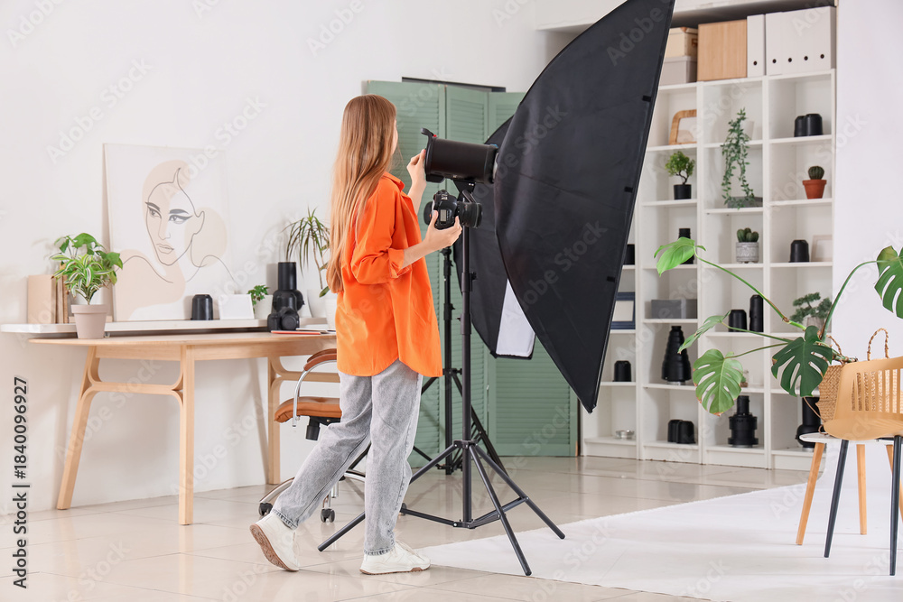 Fototapeta premium Young female photographer with houseplants, photo camera and lighting equipment preparing scene for shooting in studio