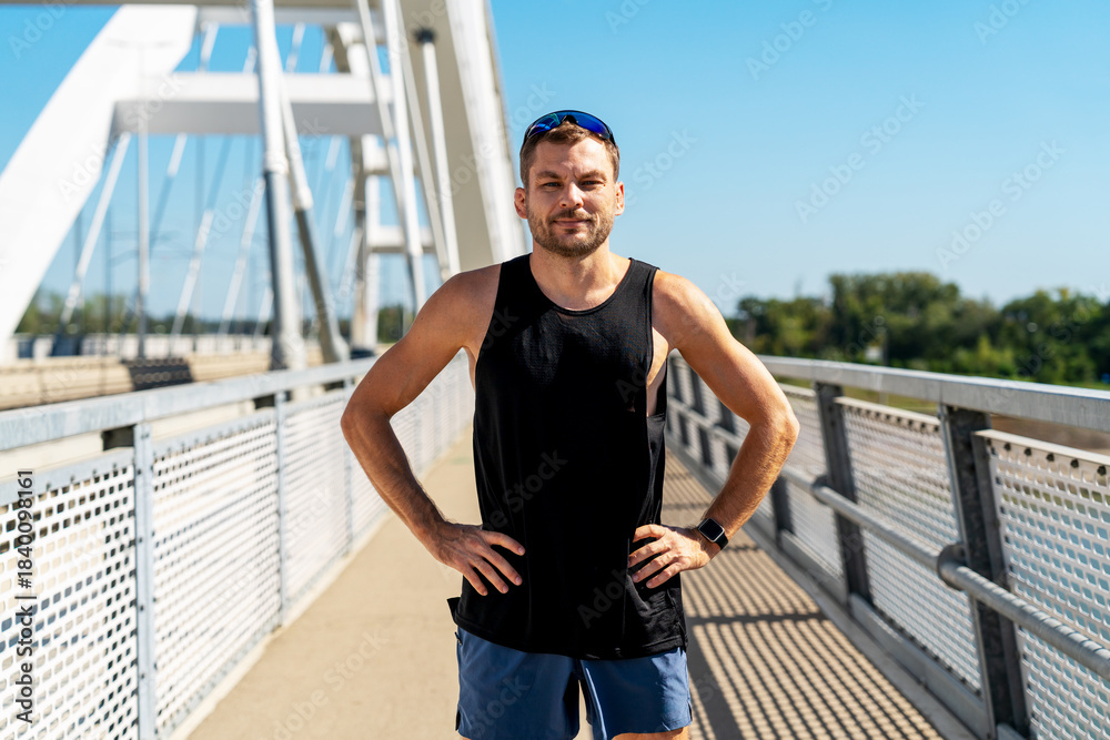 Fototapeta premium Portrait of male jogger wearing sportswear posing on urban bridge in summer.