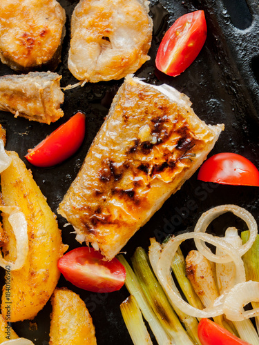 Oven pan with sliced baked potatoes, fish steaks and vegetables with onion on table.