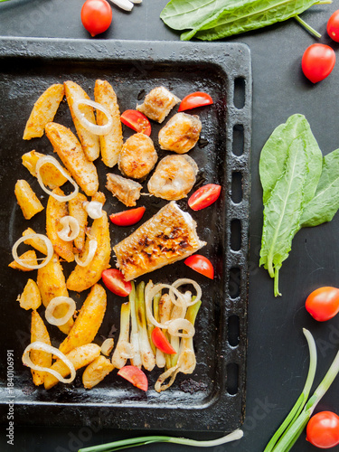 Oven pan with sliced baked potatoes, fish steaks and vegetables with onion on table.