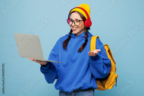 Young happy smart IT woman student wear casual clothes yellow backpack bag headphones hold books use work on laptop pc computer isolated on plain blue color background. High college lifestyle concept.