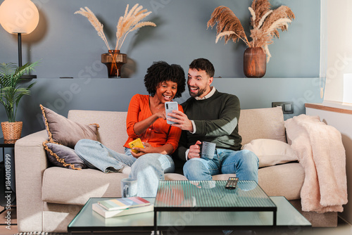 Happy couple enjoying a playful moment together as they share laughter while using their smartphone on a cozy couch, making memories and connecting through their favorite mobile applications.
