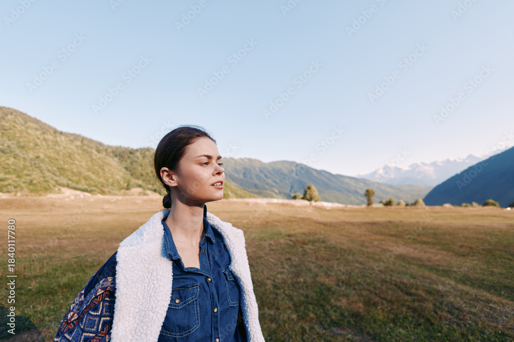 Fototapeta premium Woman in denim and shearling vest with backpack stands in a sunlit meadow near mountains, portrait with eyes closed enjoying nature, outdoor travel and calm landscape.
