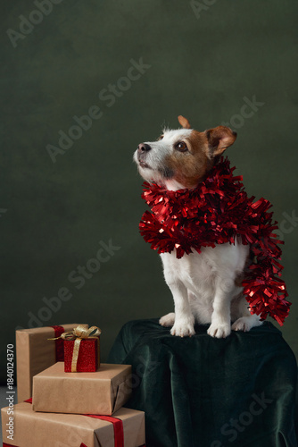A Jack Russell Terrier with a shiny red garland around its neck sits atop a pedestal next to holiday boxes. The deep green background adds contrast to the festive elements.