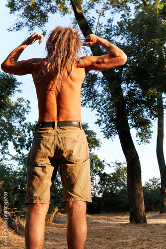 Shirtless man flexing muscles outdoors in warm sunset light surrounded by nature