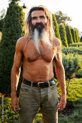 Shirtless mature man with long hair and beard standing outdoors in warm sunlight