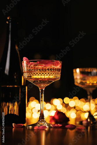A romantic setup featuring a champagne glass with rose petals, a bottle, and glowing lights, perfect for love and passion.