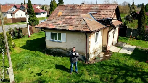 Man standing in front of a small dilapidated house in a rural setting.