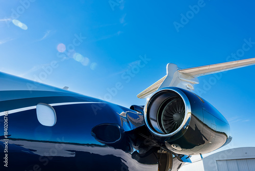 Close-up side view of a corporate jet showing the turbofan engine, fuselage, and, tail with visible lens flare and T-tail, taken in bright daylight under clear blue sky.