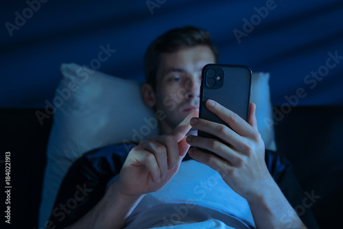 Bilde på lerret Sleepless young man browsing on his phone, doom scrolling in bed late at night lit by glowing blue screen light
