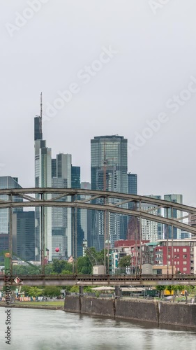 Aerial timelapse of Frankfurt's skyline through the arch of Deutschherrnbrucke (Railway Bridge), with floating boats on the Main River and water reflections. Cloudy sky frames the scene. Germany