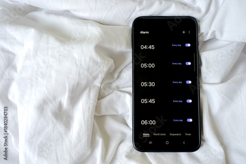Bild auf Leinwand Overhead view of a smartphone displaying multiple alarms on a rumpled white sheet or blanket