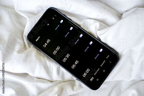 Overhead view of a smartphone displaying multiple alarms on a rumpled white sheet or blanket.