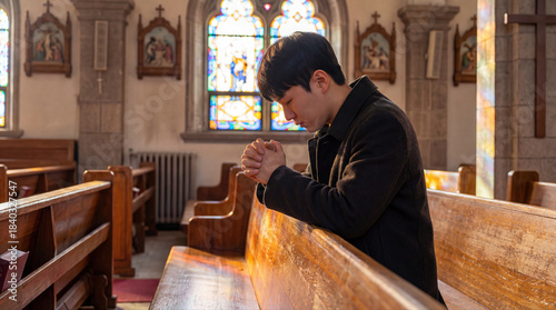 Young Asian man praying with clasped hands in a church pew. Christian faith and spirituality concept symbolizing devotion, repentance and peaceful worship inside a cathedral.