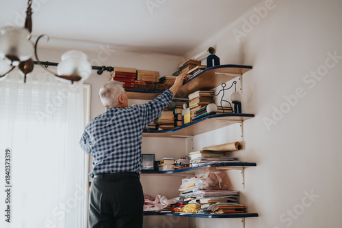 Photos An elderly man stands at a cluttered wall bookshelf, reaching for a book