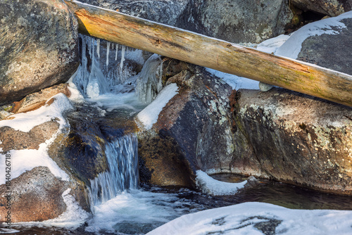 A scenic winter creek flows over rock formations, highlighted by snow and a fallen log, creating a picturesque natural landscape. High Tatras National Park, Slovakia, Europe.
