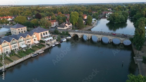 Elegant waterfront homes and a stone bridge overlook a serene river dotted with boats and waterfowl, framed by lush trees and suburban architecture.