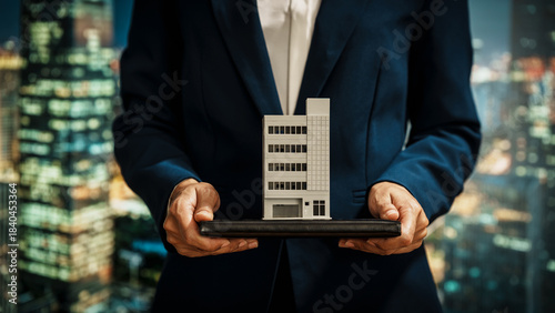 Businesswoman presents modern building model against a vibrant cityscape, real estate investment, property development, strategic planning, corporate decision-making in the commercial property sector