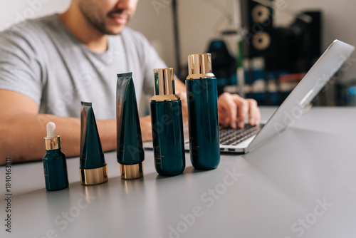 Close-up cropped shot of male hands typing on laptop amid blue and gold cosmetic bottles, tubes and droppers, working on e-commerce listings and brand management for online beauty retail.