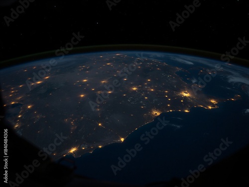 A view from space shows a city at night, with bright lights scattered across the land. The dark sky contrasts with the glow of streets and buildings below.
