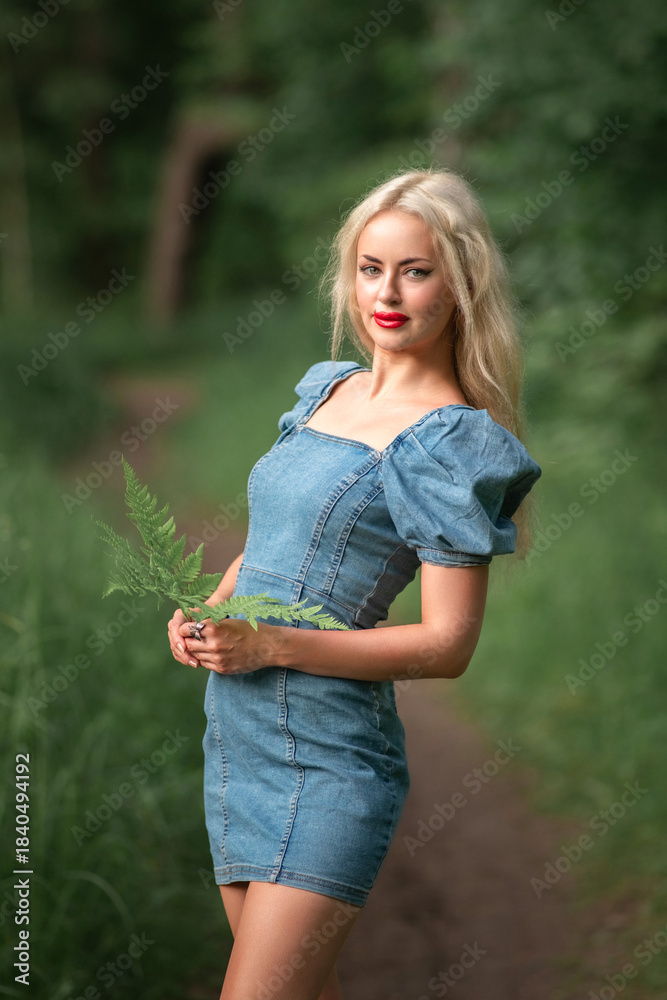 Fototapeta premium Young woman with long blonde hair wearing a denim dress stands on a path in a lush green forest, holding a fern, embodying natural beauty and serenity in nature