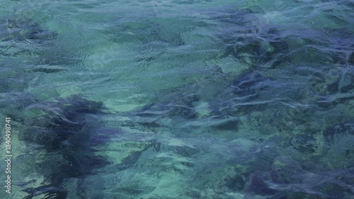 Close-up view of clear blue-green ocean water with gentle ripples and underwater rocks visible. Slow motion.