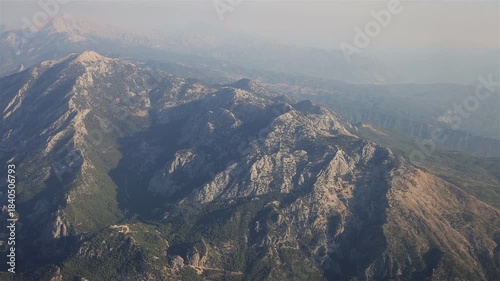 Aerial view of rugged mountain peaks and valleys covered with green vegetation under a hazy sky.