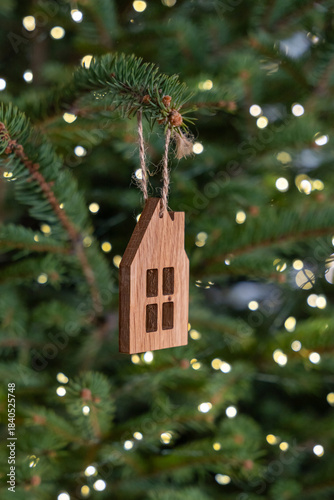 Close up of wooden house ornament hanging from fir branch of christmas tree decorated with warm fairy lights creating soft bokeh and cozy atmosphere