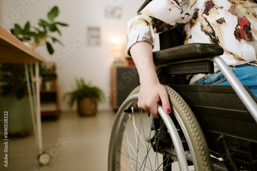 Caucasian young adult woman with disability sitting in wheelchair indoors, hand gripping wheel, partial body visible, demonstrating independence and mobility in daily life