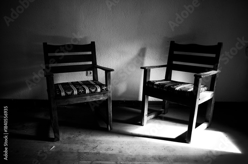 Φωτογραφία Two empty wooden chairs in a dark room with a patch of sunlight on the floor, co