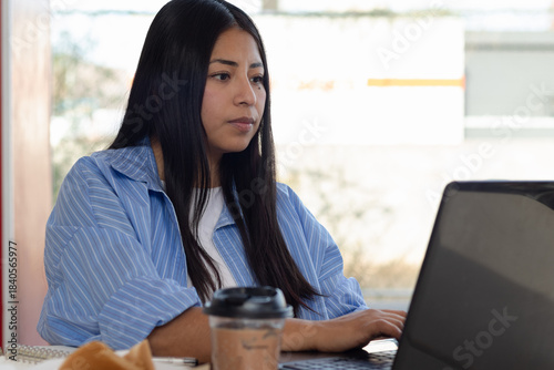 Young Mexican woman focused on her laptop screen, working efficiently from home or a remote location, with a cup of coffee and a croissant near the table. Free Download Young Mexican woman focused on her laptop screen, working efficiently from home or a remote location, with a cup of coffee and a croissant near the table. - GFXLoad