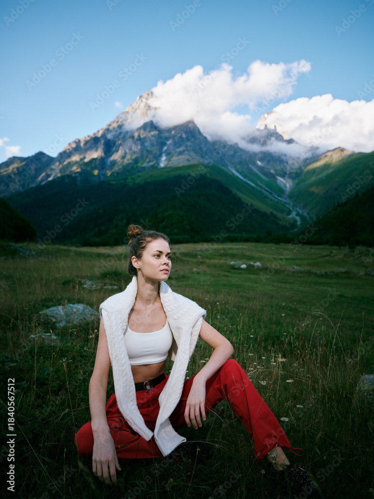 Naklejka premium Outdoor scene with a woman seated in a grassy meadow, mountains above and clouds in a clear blue sky, casual athletic wear, relaxed pose, nature and solitude in natural light and open space