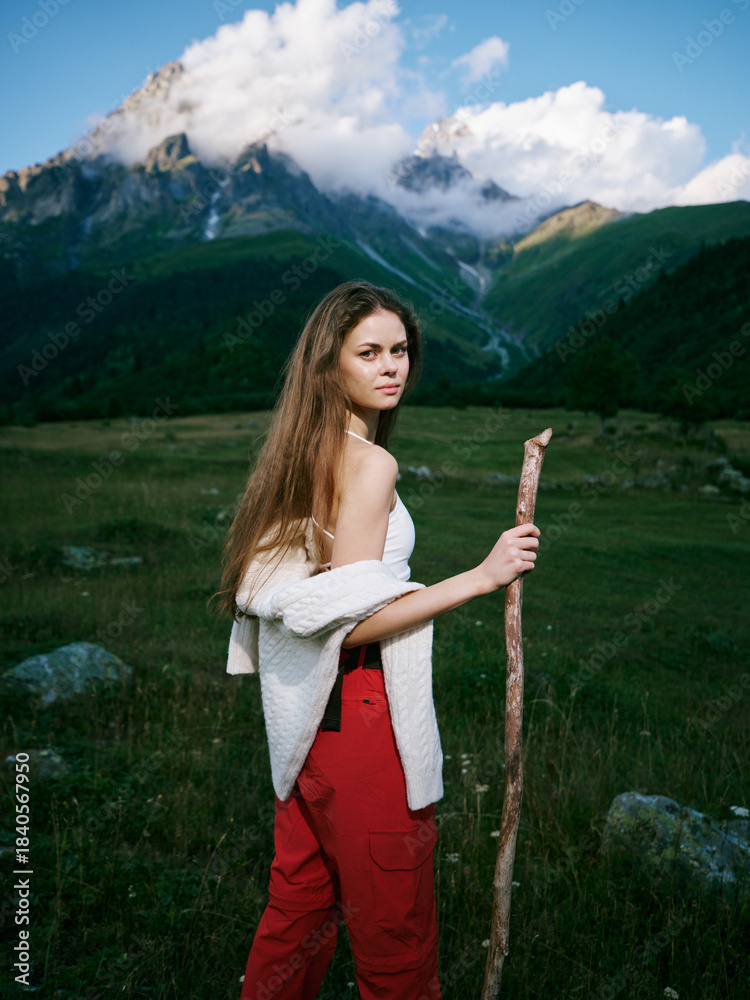 Naklejka premium Woman with a wooden staff stands in a green meadow beneath towering mountains, wearing a white cardigan and red pants, ready for a trek through pristine alpine scenery
