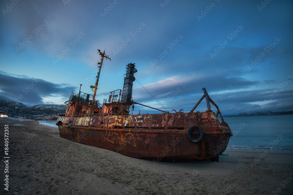 Fototapeta premium Wrecked ship rests on the shore of Teriberka in Russia during dusk with clouds above and mountains in the distance