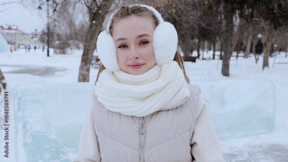 Obraz premium Bundled up in warm winter clothes, a young woman smilingly enjoys the serene snowy landscape, with cozy earmuffs and a stylish scarf providing comfort against the cold