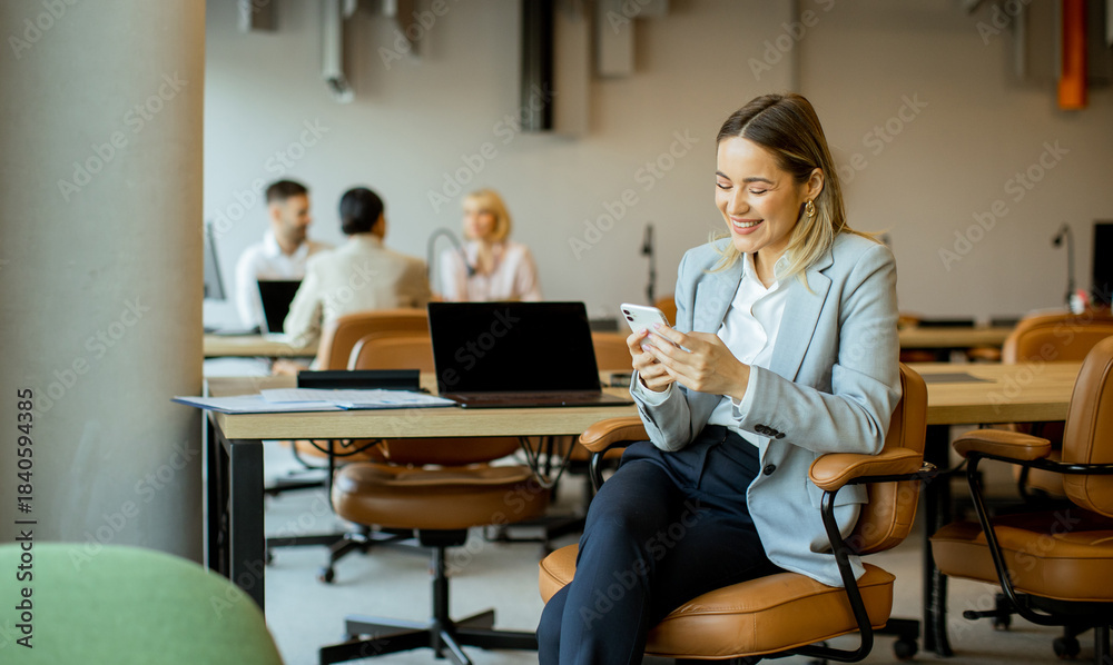 Fototapeta premium Young professional enjoys her smartphone in a lively office setting