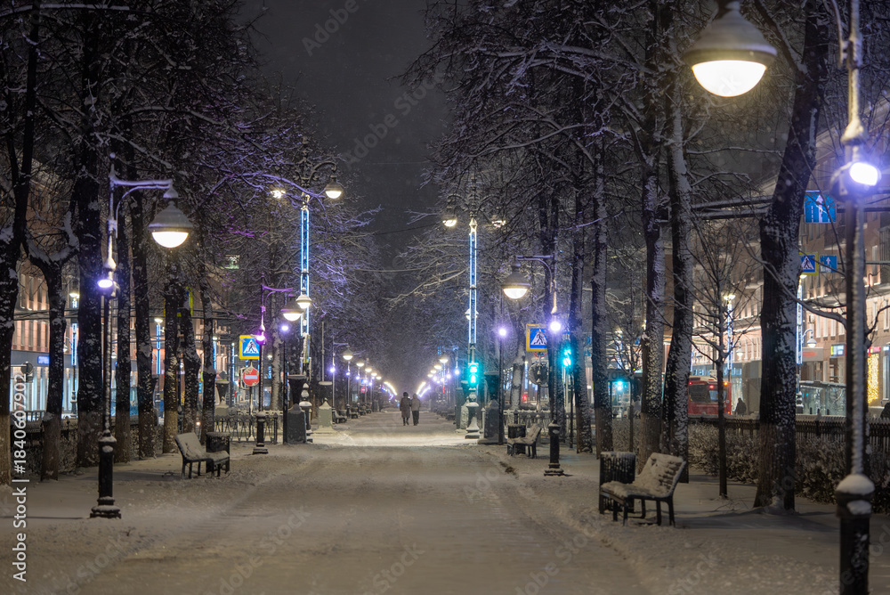 Fototapeta premium Snow-covered boulevard with streetlights and bare trees at night, creating a peaceful winter scene as people walk along the illuminated path in the city