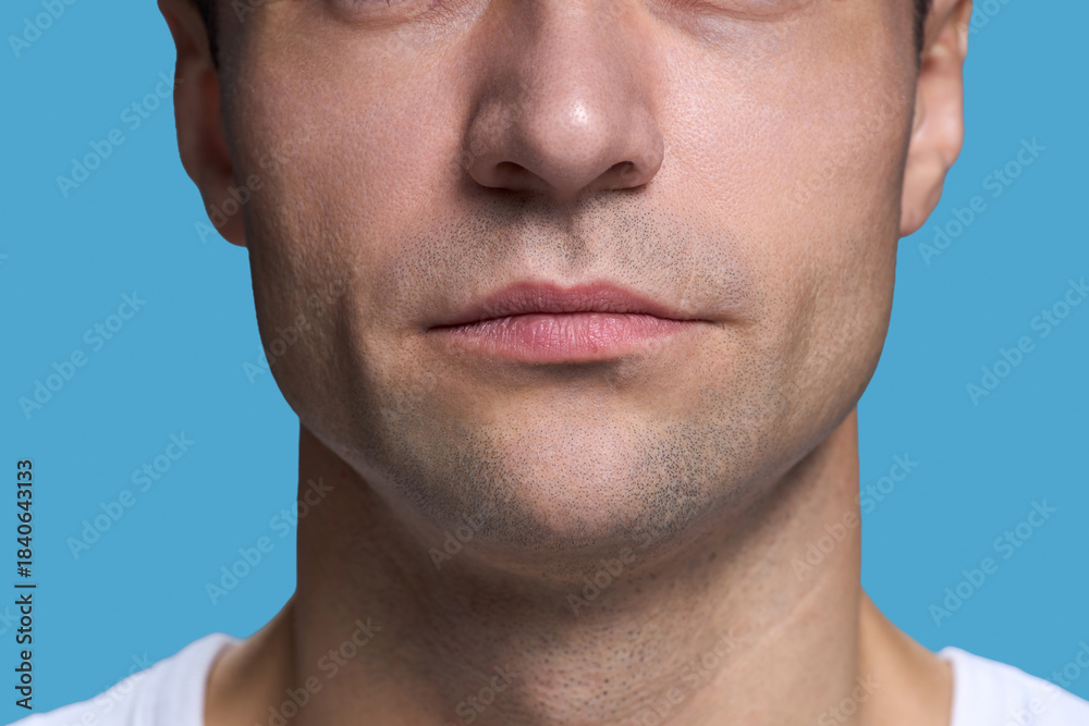 Obraz premium Caucasian young adult man showing lower half of face with neutral expression against blue background, lips closed, stubble visible, wearing casual shirt